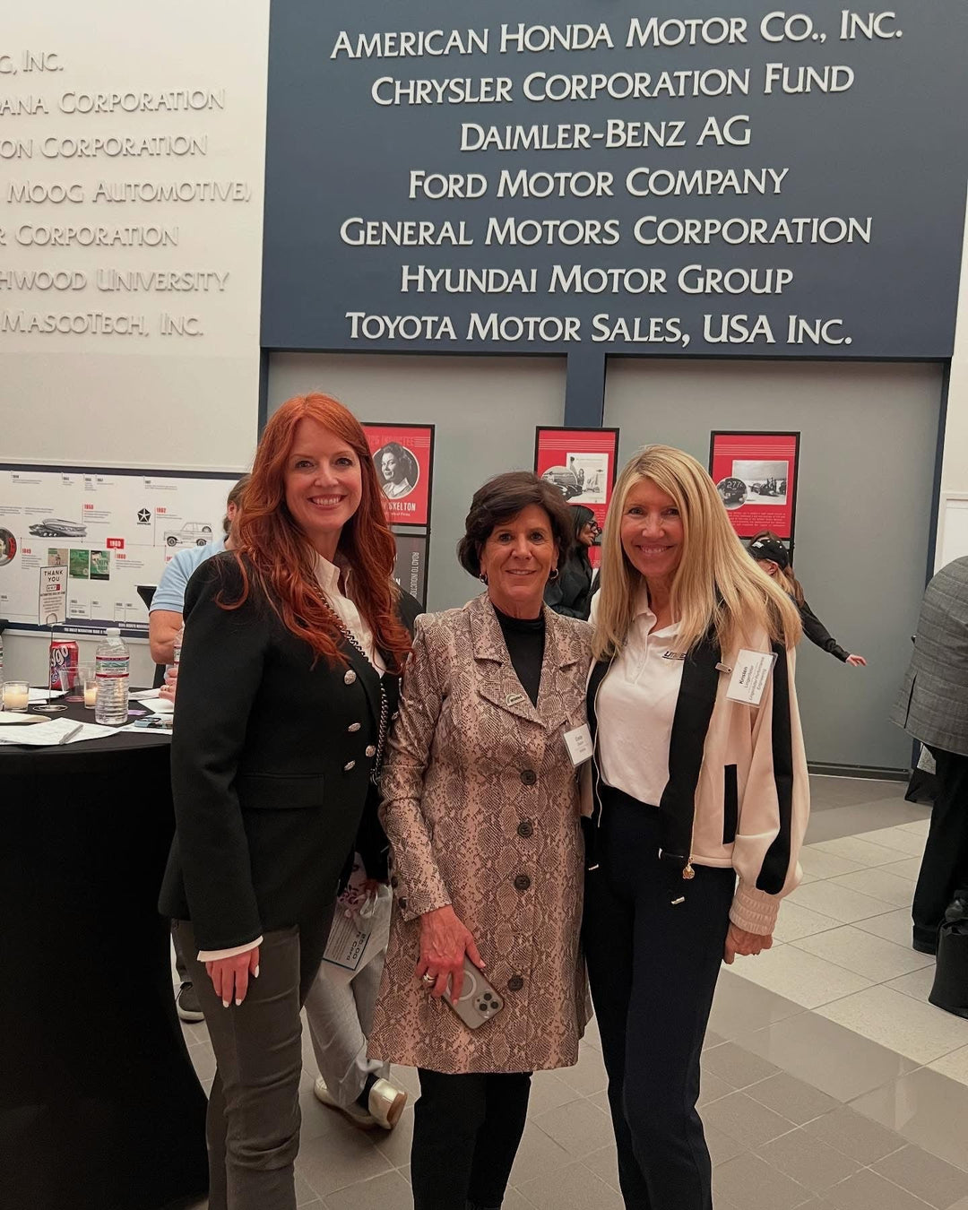 Three women pose together at a motorsport event in Detroit, surrounded by auto company logos.